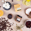 Upcircle Face Scrub Citrus Blend laid on a table with its incorporating raw ingredients on show around the jar, box and another jar with the product visible.