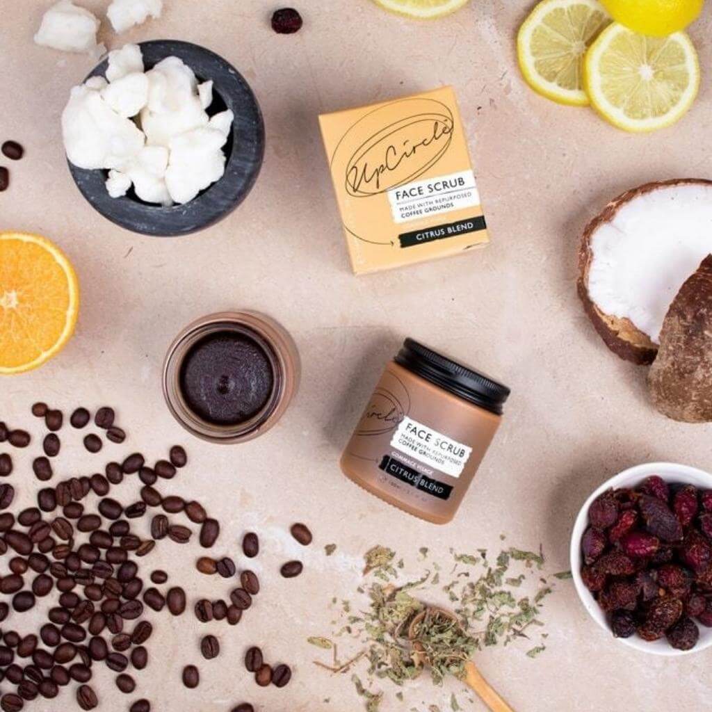 Upcircle Face Scrub Citrus Blend laid on a table with its incorporating raw ingredients on show around the jar, box and another jar with the product visible.