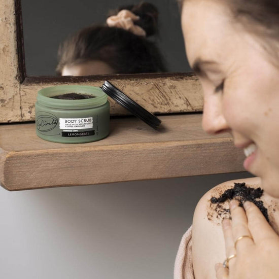 Upcircle Body Scrub with Lemongrass jar with lid off displayed on a mirror and shelf, with a model applying the Body Scrub to her shoulder.