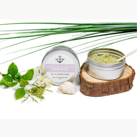 The Salt Parlour Jasmine & Matcha Green Tea REPAIR salt and sulphur scrub. A desktop view with the yellow-green scrub on show with a wooden spatula in it. The tin is on a wood coaster and a green palm frond in the background.