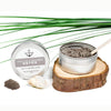 The Salt Parlour Black Clay and Vanilla Detox salt and sulphur scrub. A desktop view with the dark scrub on show with a wooden spatula in it. The tin is on a wood coaster and a green palm frond in the background.