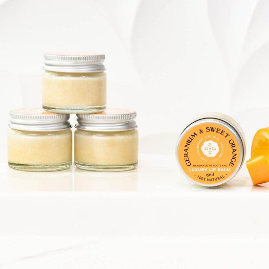 The Edinburgh Natural Skincare Co. Geranium and Sweet Orange Luxury Lip Balm. Three jars stacked in a pyramid, and a jar on its side with an slither of orange peel beside it.