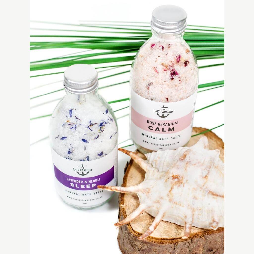 The Salt Parlour - Lavender and Neroli SLEEP Mineral Bath Salts. - Rose Geranium CALM Mineral Bath Salts. With palm fronds in the background and a conch shell on a wooden coaster in the front.