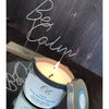 Orli Calm Therapy Massage Candle in Aluminium tin lifestyle image. Candle lit and ready to be used as a massaging oil. Be Calm in the background.