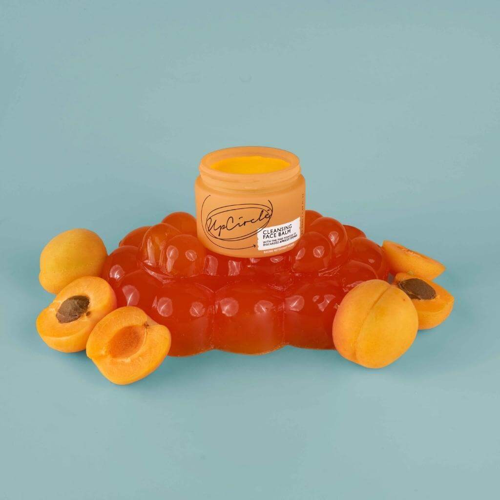 Upcircle Cleansing Face Balm with its lid on a jellied mound of apricots and fresh halved apricots around the mound. With a blue background.