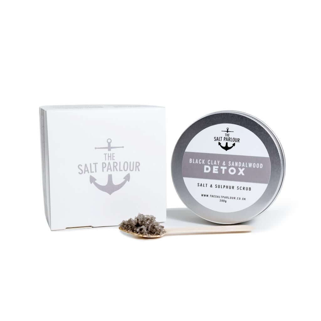 The Salt Parlour Black Clay and Vanilla DETOX salt and sulphur scrub with wooden spoon holding a small amount of salt scrub. The box and tin are upright on a white background.
