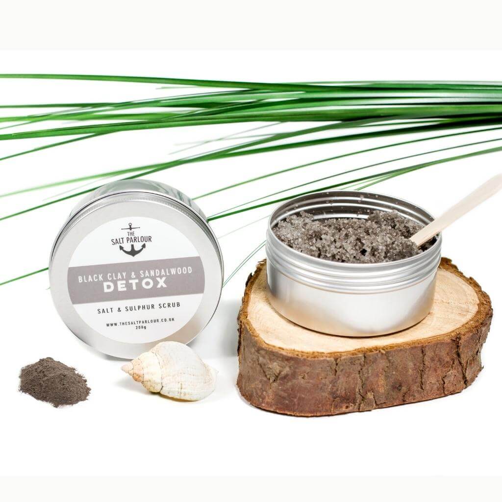 The Salt Parlour Black Clay and Vanilla Detox salt and sulphur scrub. A desktop view with the dark scrub on show with a wooden spatula in it. The tin is on a wood coaster and a green palm frond in the background.
