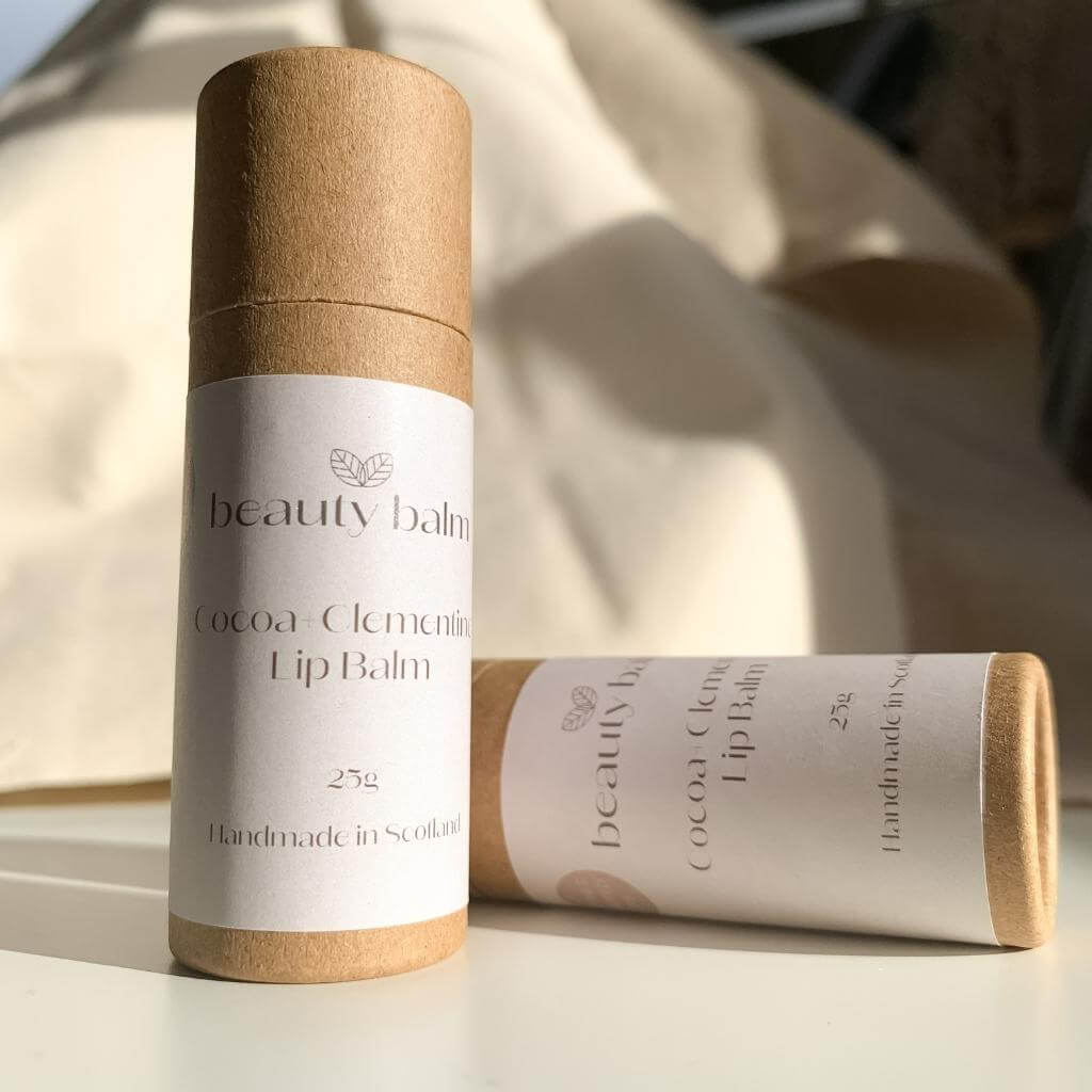 Beauty Balm Cocoa and Clementine Lip Balm Sticks in Cardboard Tubes. Two tubes laying on their side on a white table.