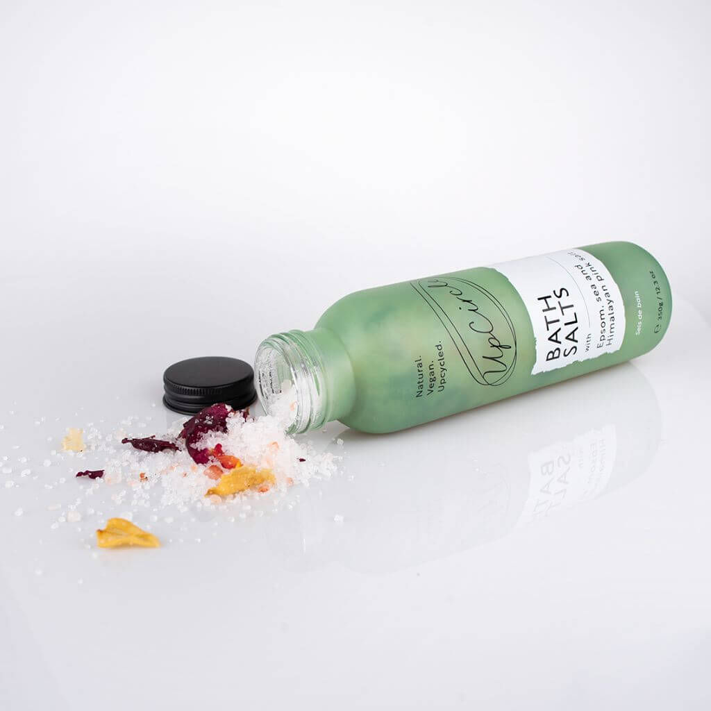UpCircle Bath Salts with Epsom, Sea and Himalayan Pink Salt, and Rose Petals. Natural, sustainable, vegan & cruelty-free. . Laying on the side with Salts and Petals visible with Glass Bottle and Aluminium Lid on a white background.